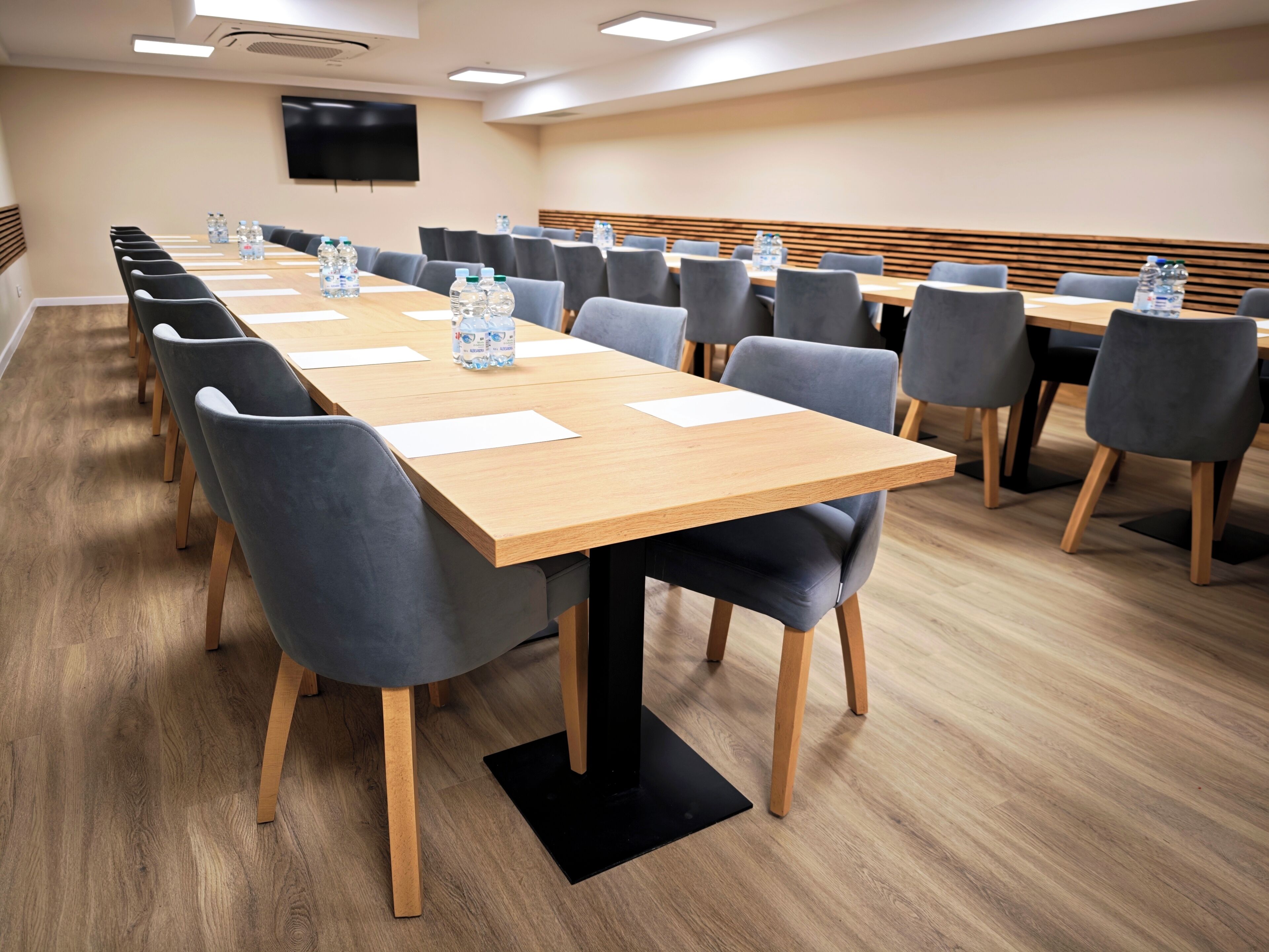 A conference room with long tables, chairs, water bottles, and a flat-screen TV on the wall.