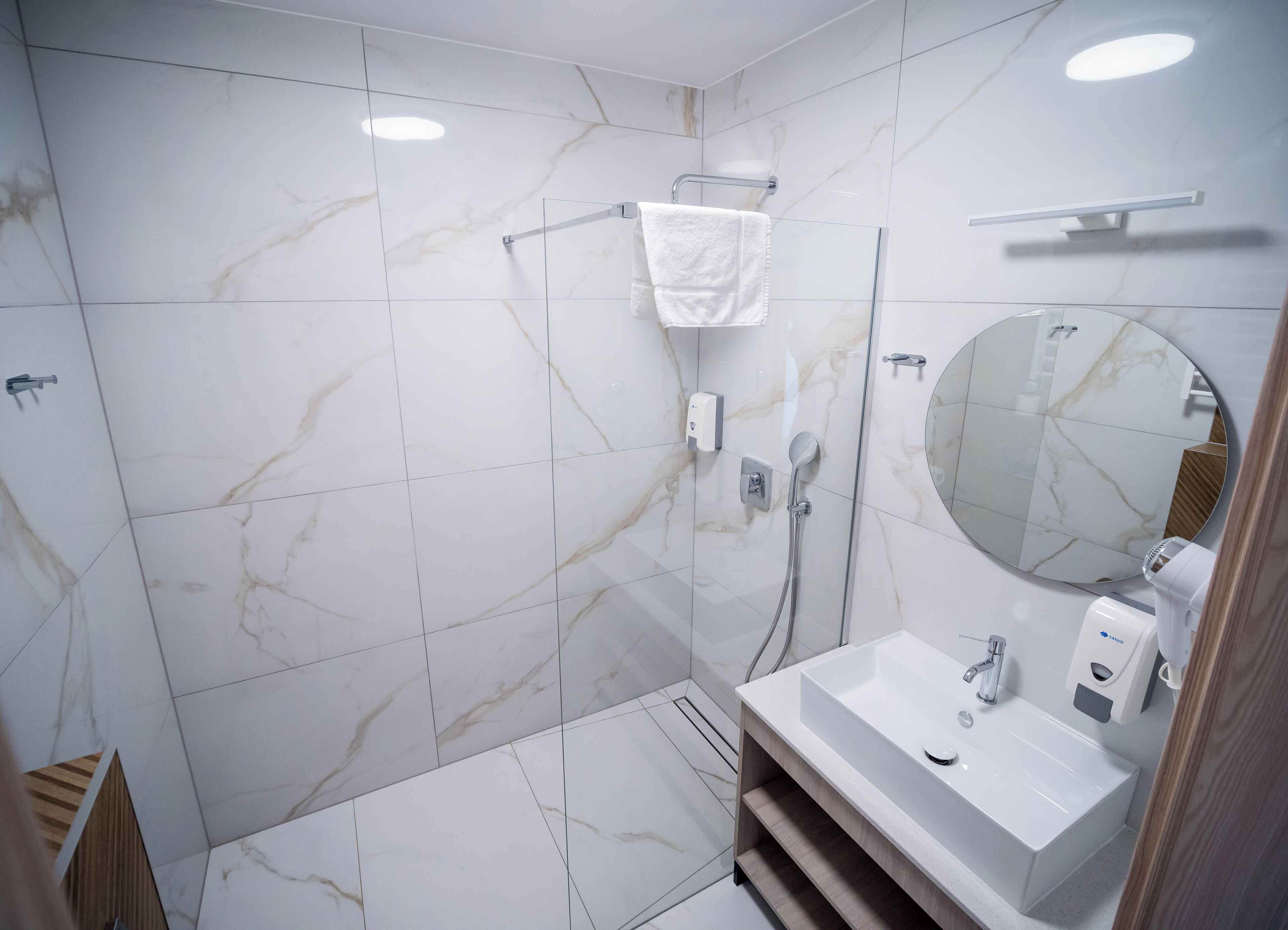 A modern bathroom with marble tiles, a round mirror, a white sink, and a handheld showerhead.