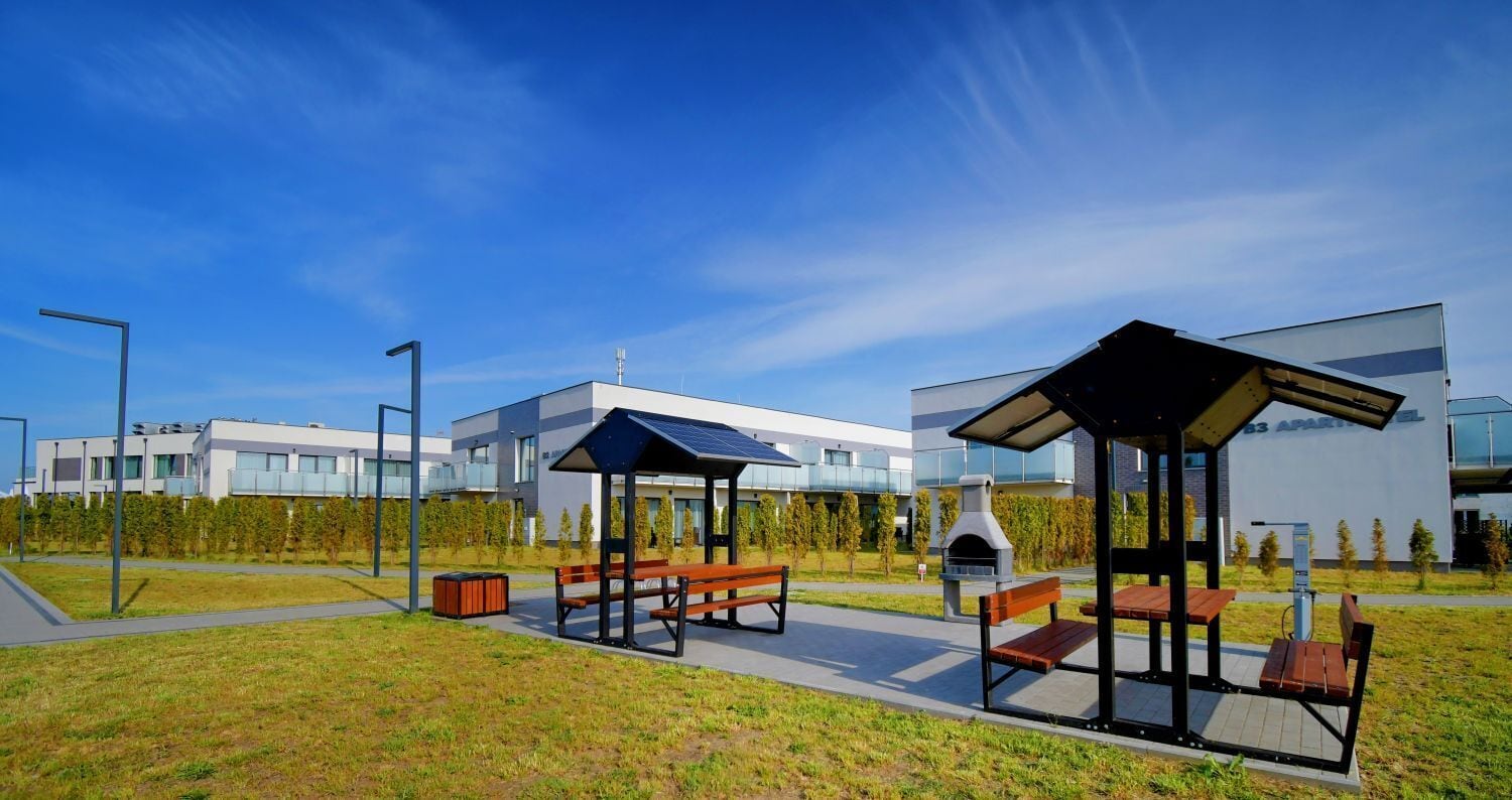 A modern outdoor seating area with benches and a barbecue grill, set against a backdrop of contemporary buildings and a clear sky.