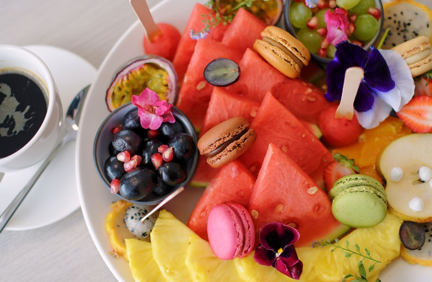 A plate of assorted fruits and macarons with a cup of coffee on the side.