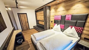 Ett modernt hotellrum med en stor säng, en träpanel vid sänggaveln med rosa och röda geometriska mönster, en inbyggd bänk och en liten sittgrupp.