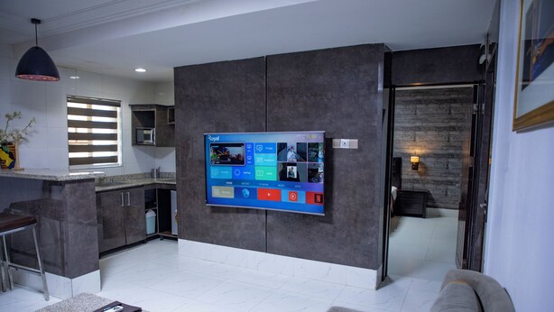 A modern living room with a flat-screen TV mounted on the wall, a grey sofa, a coffee table, and a kitchen area in the background. - NewLand Hotels and Apartments (Abuja)