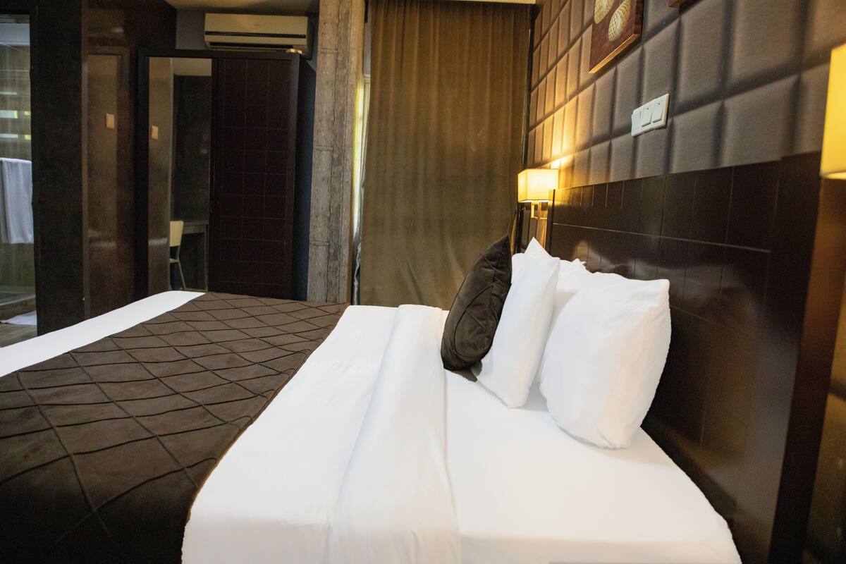 A hotel room with a large bed, a headboard with a light fixture, a dark tiled floor, and a view of the outside through a window.