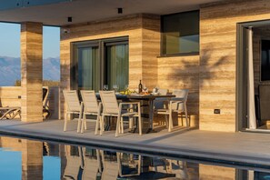 A modern outdoor dining area with a pool, featuring a table set for four, surrounded by chairs, and a view of mountains in the background. - Resort Travera (Privlaka)