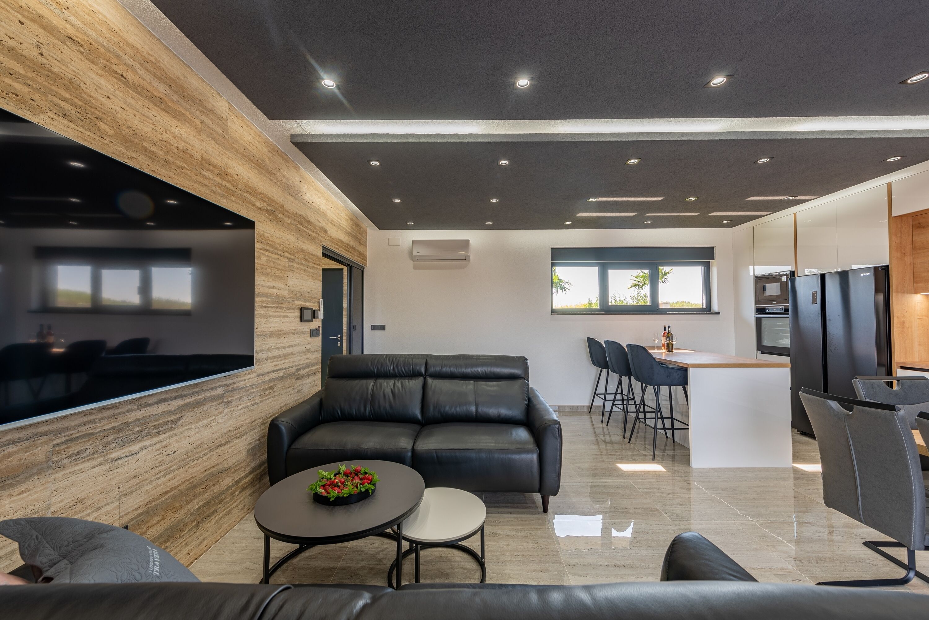 A modern living room with a black leather sofa, a small round table with a flower centerpiece, and a dining area with bar stools.