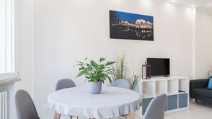 A modern living room with a round dining table, grey upholstered chairs, a potted plant, a flat-screen TV on a white stand, and a cityscape painting on the wall.