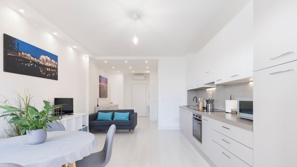 A modern kitchen with white cabinets, a dining area with a table and chairs, and a living area with a sofa and a TV.