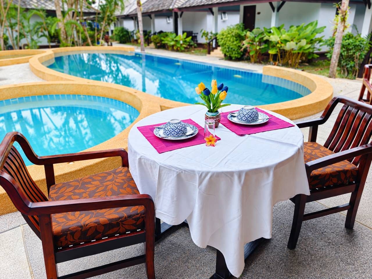 A poolside dining area with a table set for two, surrounded by chairs with patterned cushions.