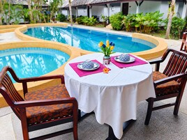 Zona de comedor junto a la piscina con una mesa preparada para dos personas, rodeada de sillas con cojines estampados.