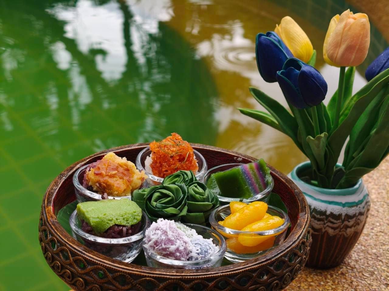 Una bandeja de postres coloridos con un tazón decorativo, un florero con tulipanes y un estanque al fondo.
