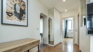 Un corridoio con pavimento in legno, un quadro appeso alla parete e una moderna zona cucina visibile attraverso una porta aperta.