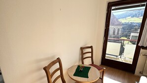 Un pequeño comedor con una mesa redonda y dos sillas de madera, vista de un edificio a través de una ventana, y suelo de madera.