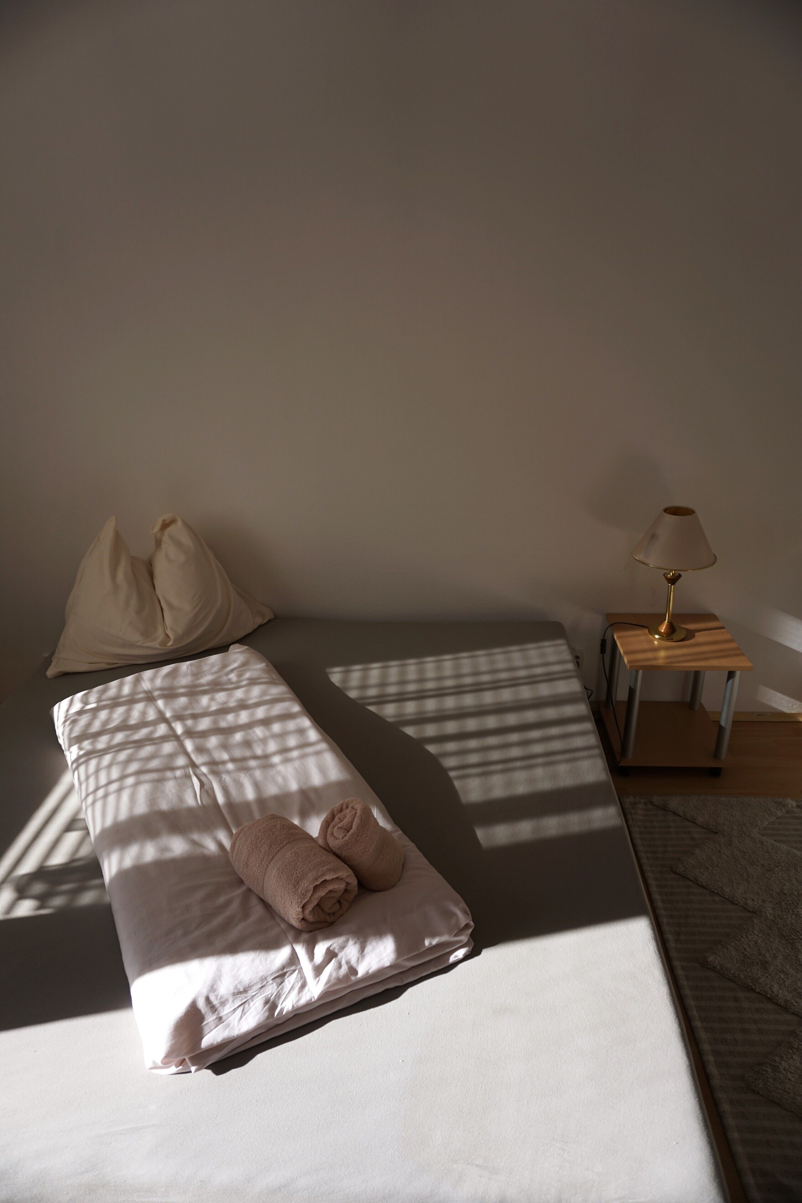 Een net opgemaakt bed met kussens en een opgerolde handdoek, een bedlamp en een klein houten nachtkastje.