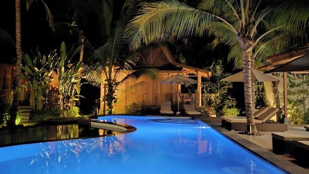 Un área de piscina nocturna con palmeras y una estructura de techo de paja.