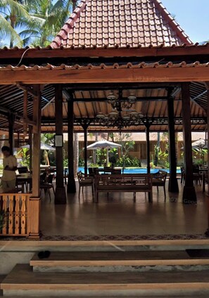 A covered outdoor seating area with wooden furniture and a pool in the background. - Villa Lova Sambirenteng (Tejakula)