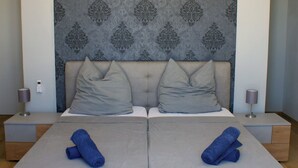 Una cama doble con dos almohadas y toallas enrolladas, una mesita de noche con lámpara, y un cabecero estampado.