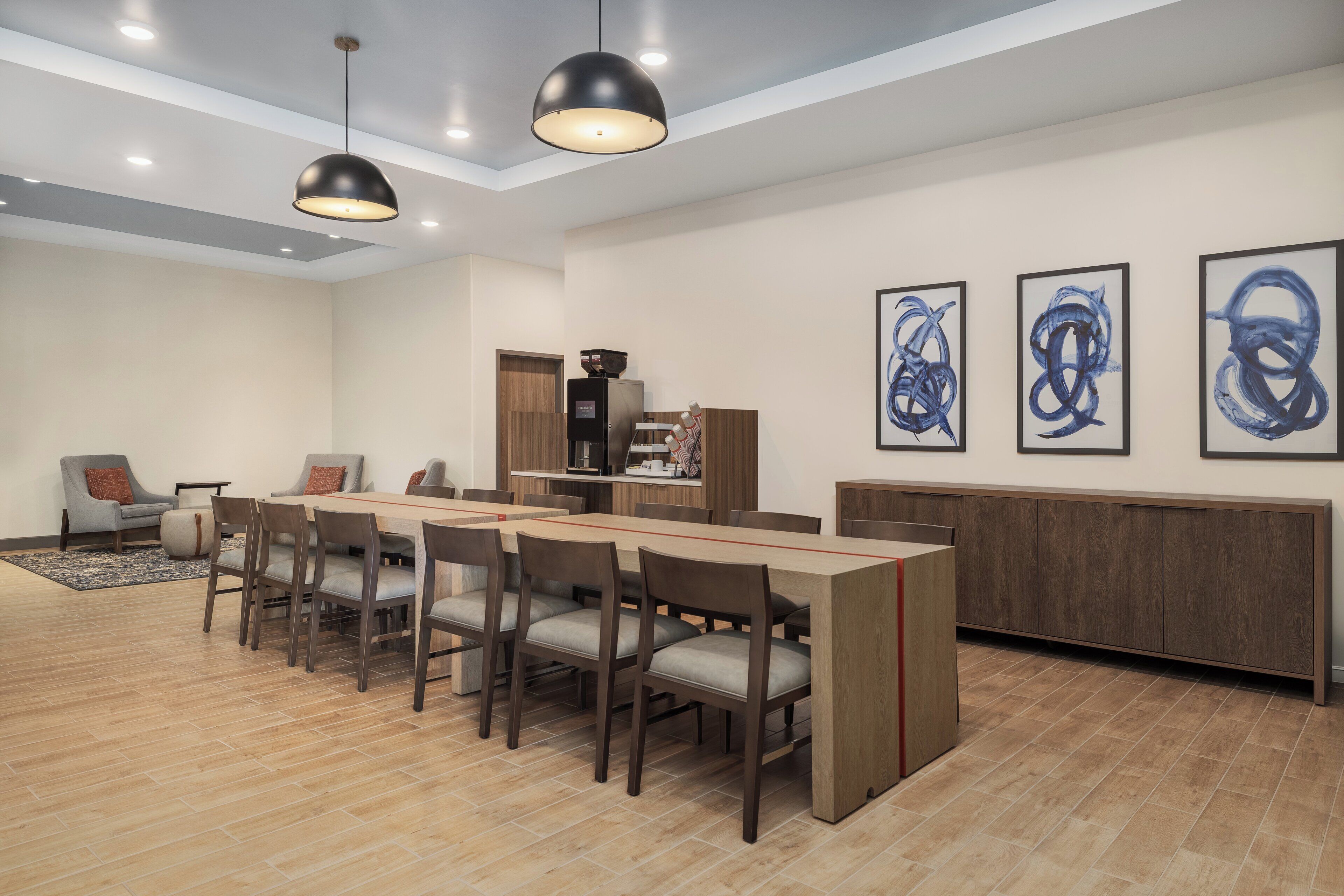 A modern dining area with a long table, chairs, pendant lighting, artwork on the wall, and a kitchen in the background.