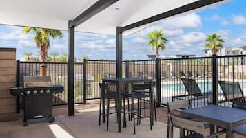 A covered outdoor patio with a grill, tables, chairs, and a view of a swimming pool.