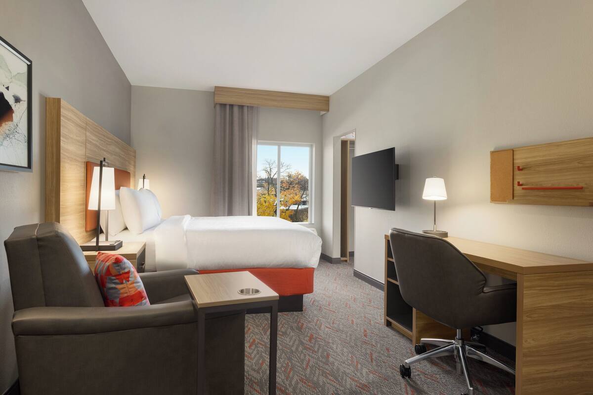 Une chambre d’hôtel équipée d’un lit, d’un bureau, d’une chaise, d’une télévision et d’une fenêtre donnant sur la verdure.