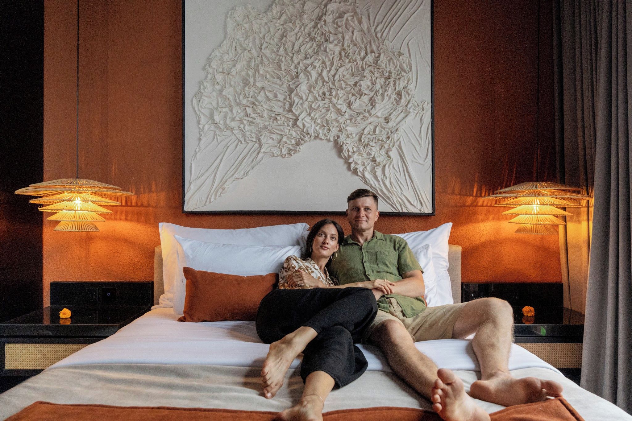 A couple sitting on a bed in a hotel room with a large artwork above them and two bedside lamps.