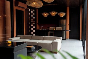 A modern living room with a sectional sofa, a coffee table, and a kitchen area with a refrigerator and hanging lights. - Earth Villa Canggu by Ini Vie Hospitality (Canggu)