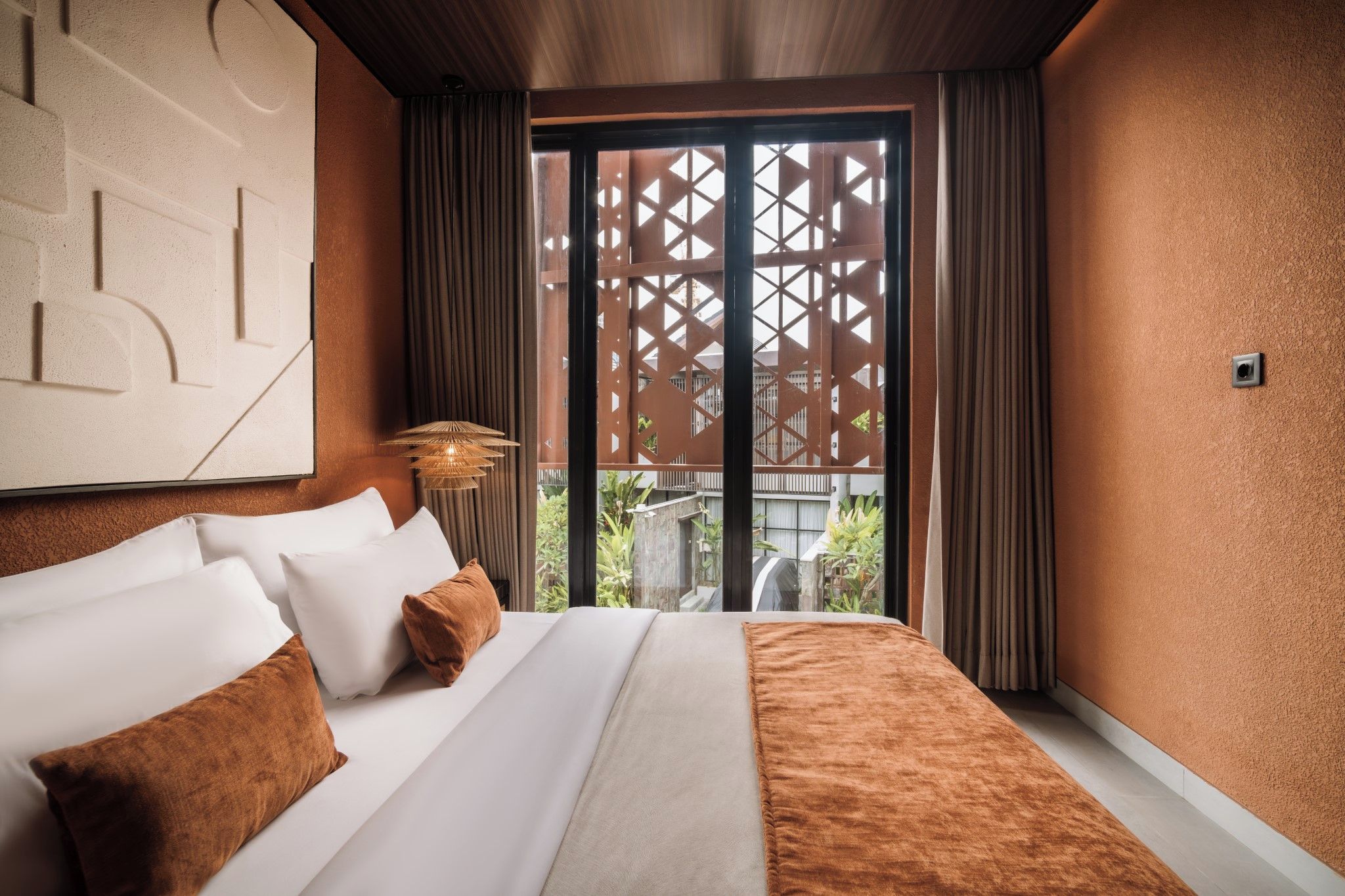A hotel room with a bed, curtains, a window with a view, and a wall-mounted light fixture.