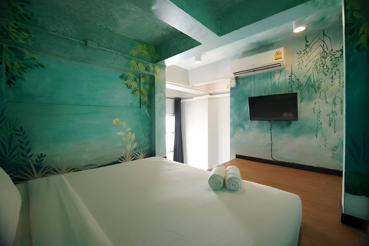 A hotel room with a bed, a flat-screen TV mounted on the wall, and a mural of greenery on the walls.