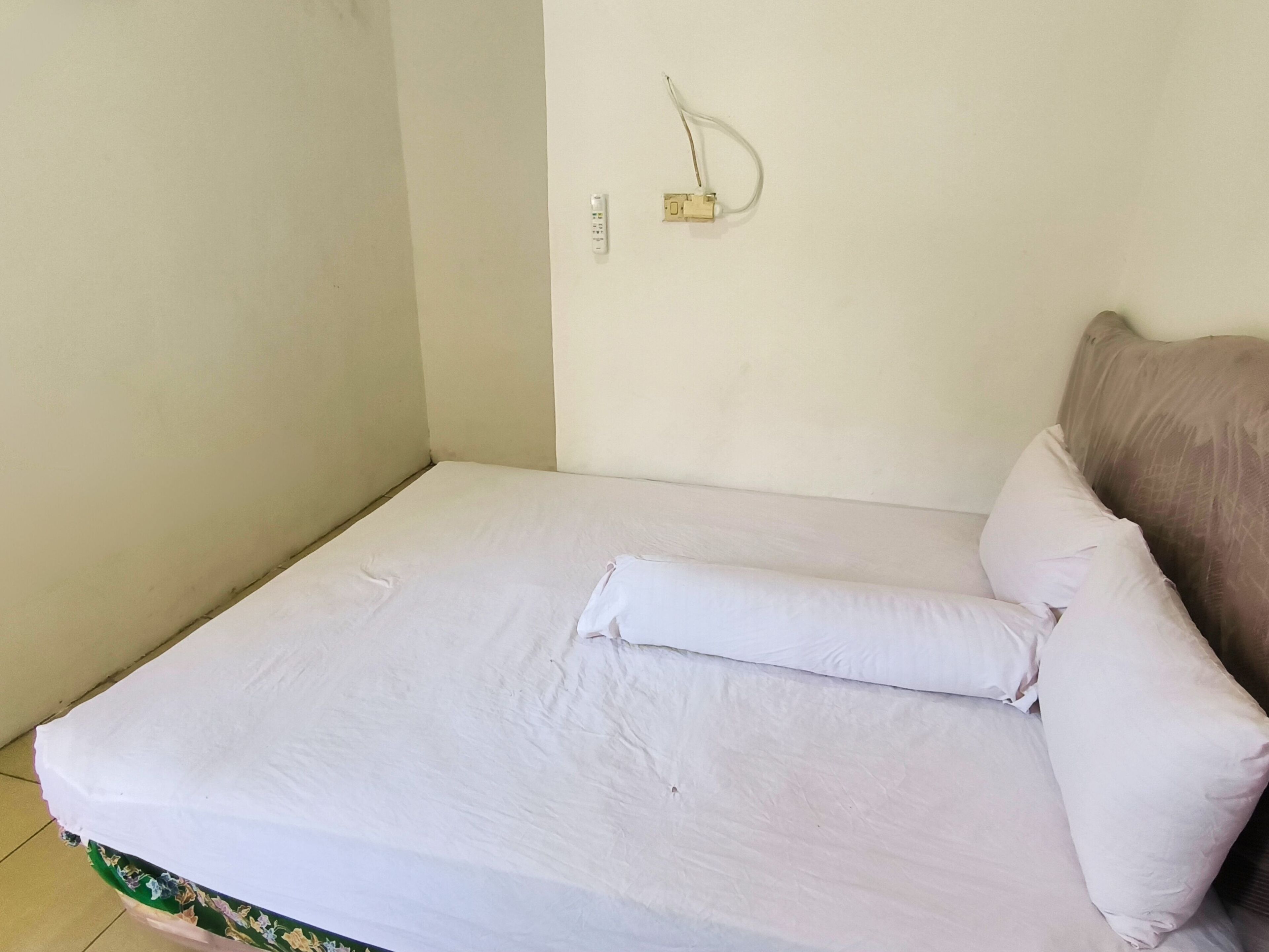 Un petit lit bien fait, avec une literie blanche et un seul oreiller, une prise électrique au mur et une tête de lit marron.