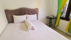Een net opgemaakt bed met witte lakens en een bruin hoofdeinde, een klein rond tafeltje met een stoel, en een raam met groene en gele gordijnen.