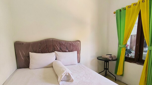 Een netjes opgemaakt bed met kussens en een gevouwen handdoek, een klein rond tafeltje met een stoel, en een raam met groene en gele gordijnen.