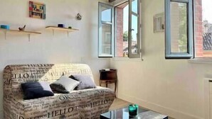 Un salon avec un canapé recouvert d’un tissu imprimé de motifs de journaux, une petite table sur laquelle est posée une bougie, et une fenêtre dont le volet est ouvert.