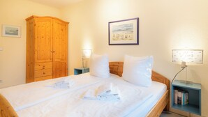 Een net opgemaakt bed met witte lakens en handdoeken, een houten garderobe, een nachtkastje met een lamp en een ingelijst schilderij aan de muur.