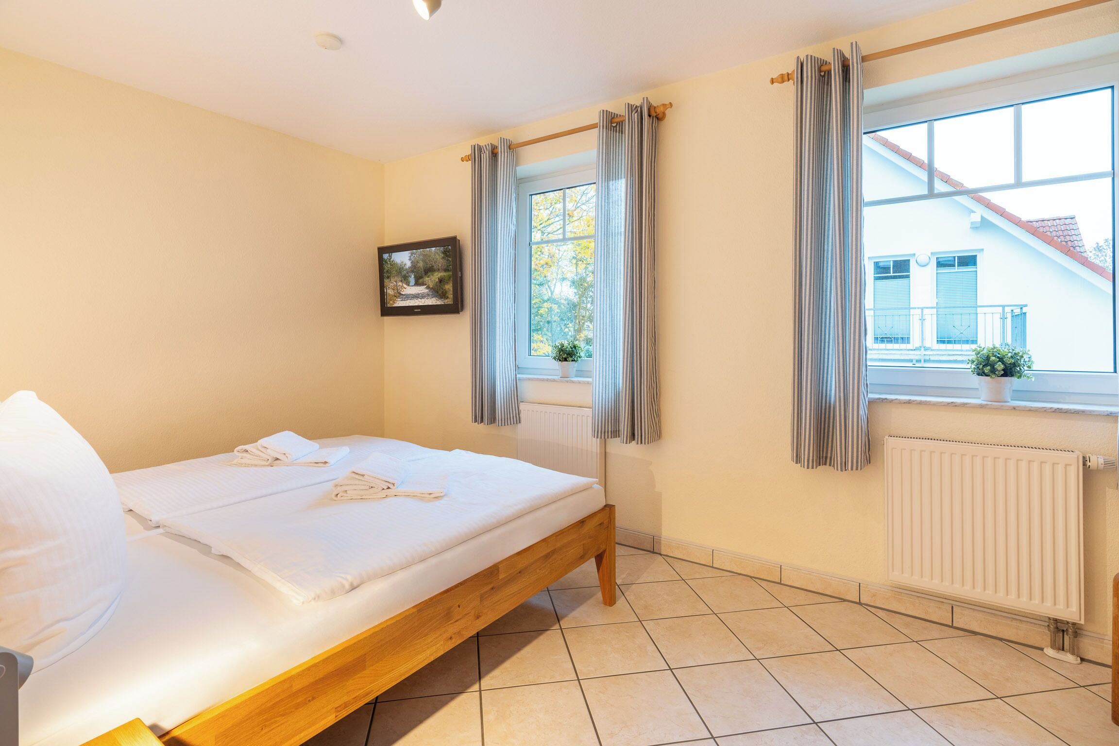 A neatly made bed with white linens, a window with curtains, a radiator, and a framed picture on the wall.