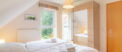 A bedroom with two beds, a large window, a wooden wardrobe, and a ceiling light fixture.