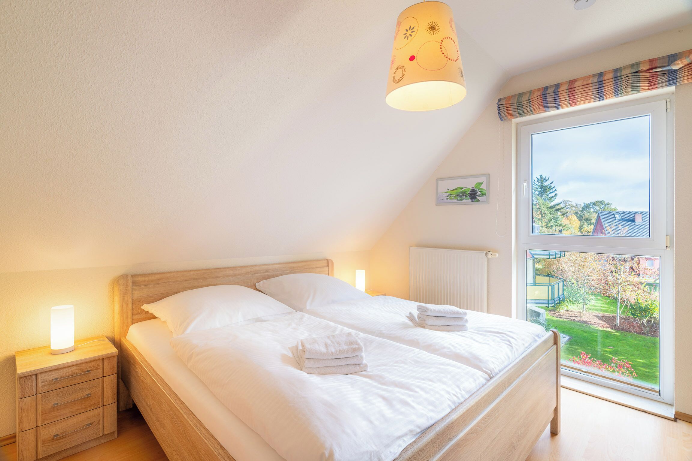 Une chambre à coucher avec un grand lit, deux tables de chevet, une fenêtre donnant sur un jardin et un lampadaire au plafond.