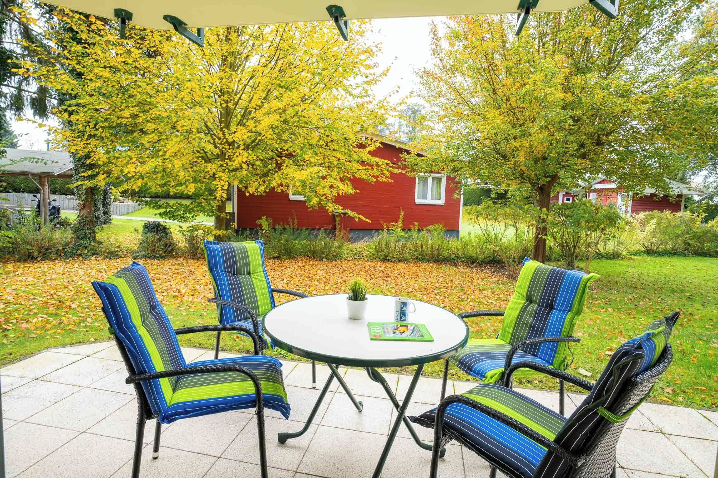 Eine Terrasse mit vier gestreiften Stühlen und einem kleinen Tisch, umgeben von einem Garten mit einem roten Haus im Hintergrund.