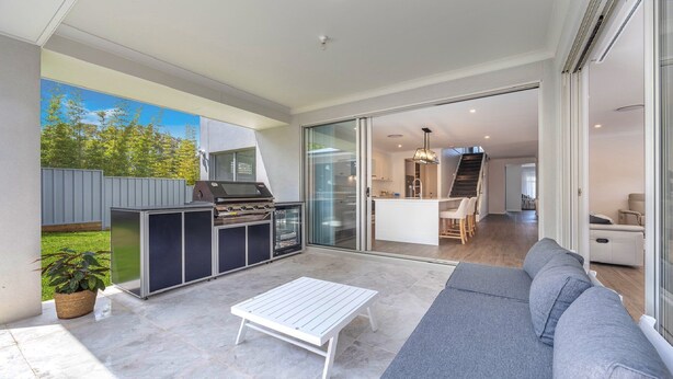 A modern outdoor patio with a barbecue, a sofa, and a glass door leading to a kitchen and living area. - Coastal Ambience by HolidayCo. (KILLCARE HEIGHTS)