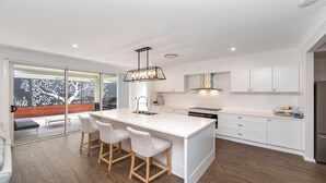 A modern kitchen with a central island, bar stools, white cabinetry, and a view of a deck with a tree. - Coastal Ambience by HolidayCo. (KILLCARE HEIGHTS)