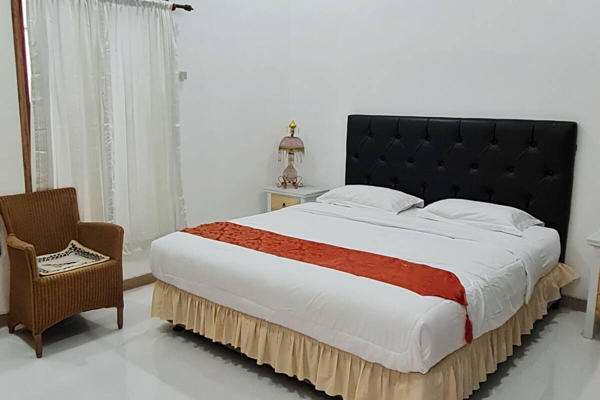 Un lit bien fait, avec une tête de lit noire, une couvre-lit blanche et un élément décoratif rouge. On y trouve une chaise en osier et une petite table de chevet avec une lampe.