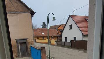Une vue à travers une fenêtre donnant sur une rue bordée d’immeubles, avec une poubelle bleue et un lampadaire.