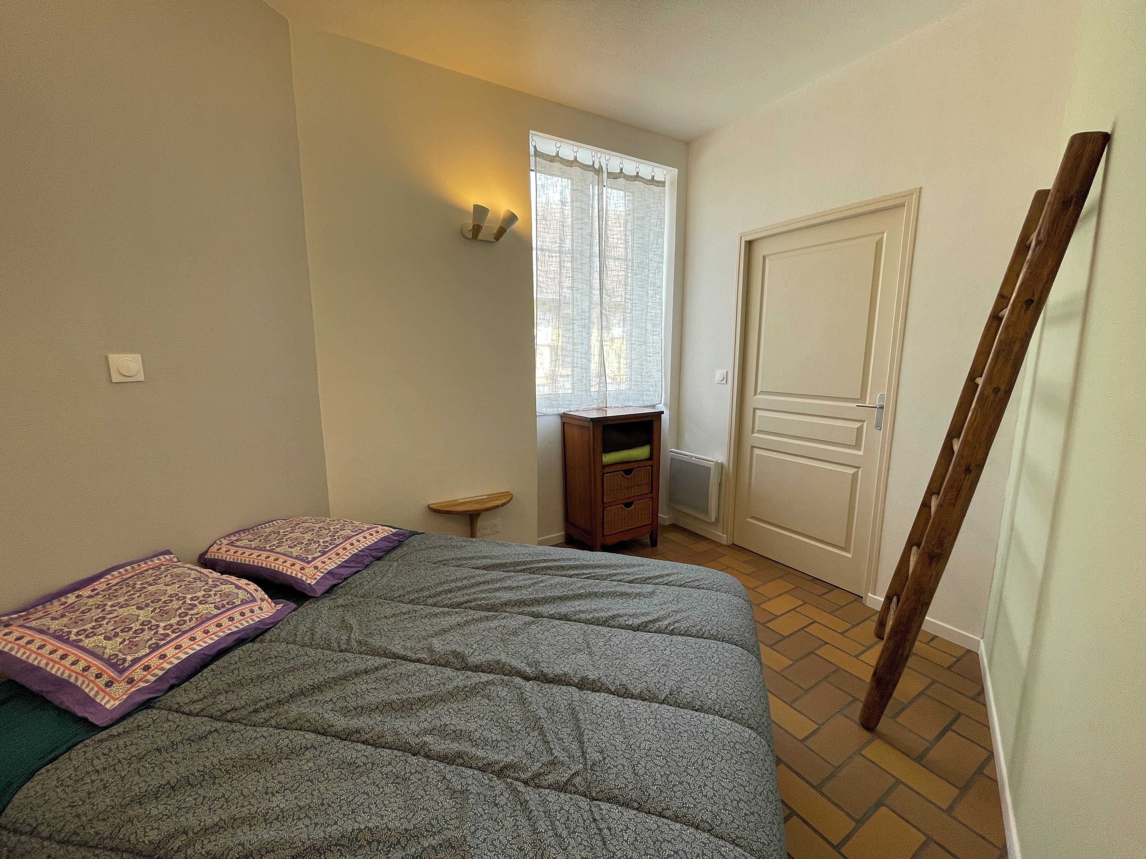 A bedroom with a bed, a small wooden nightstand, a window with sheer curtains, a radiator, and a door.