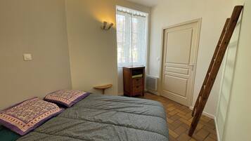 A bedroom with a bed, a small wooden nightstand, a window with sheer curtains, a radiator, and a door.