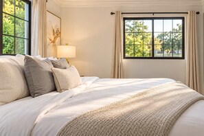 A neatly made bed with a white comforter and several pillows, a bedside lamp, and a window with curtains. - The Sundancer Oasis|Old Town|Pool+Spa|Rooftop Deck (Scottsdale)