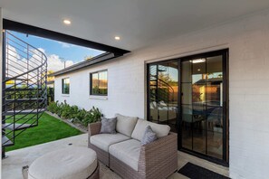 A modern outdoor living area with a sofa, a round ottoman, a spiral staircase, and a glass door leading to a patio.