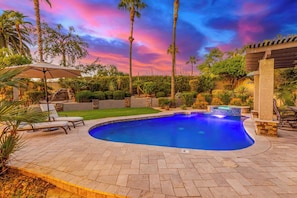 A swimming pool with a herringbone tile pattern, surrounded by palm trees and a stone wall.