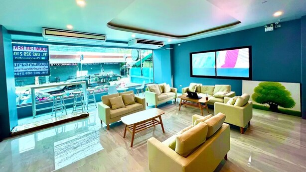 A modern hotel lobby with seating areas, a large window with a view of a busy street, and air conditioning units on the ceiling. - THE NICE  (Patong)