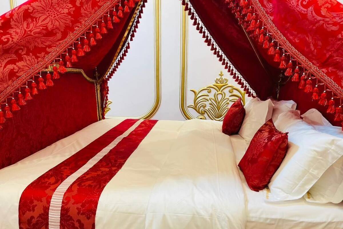 Un lit avec une literie rouge et blanche, une tête de lit dorée et des glands rouges sur les rideaux.