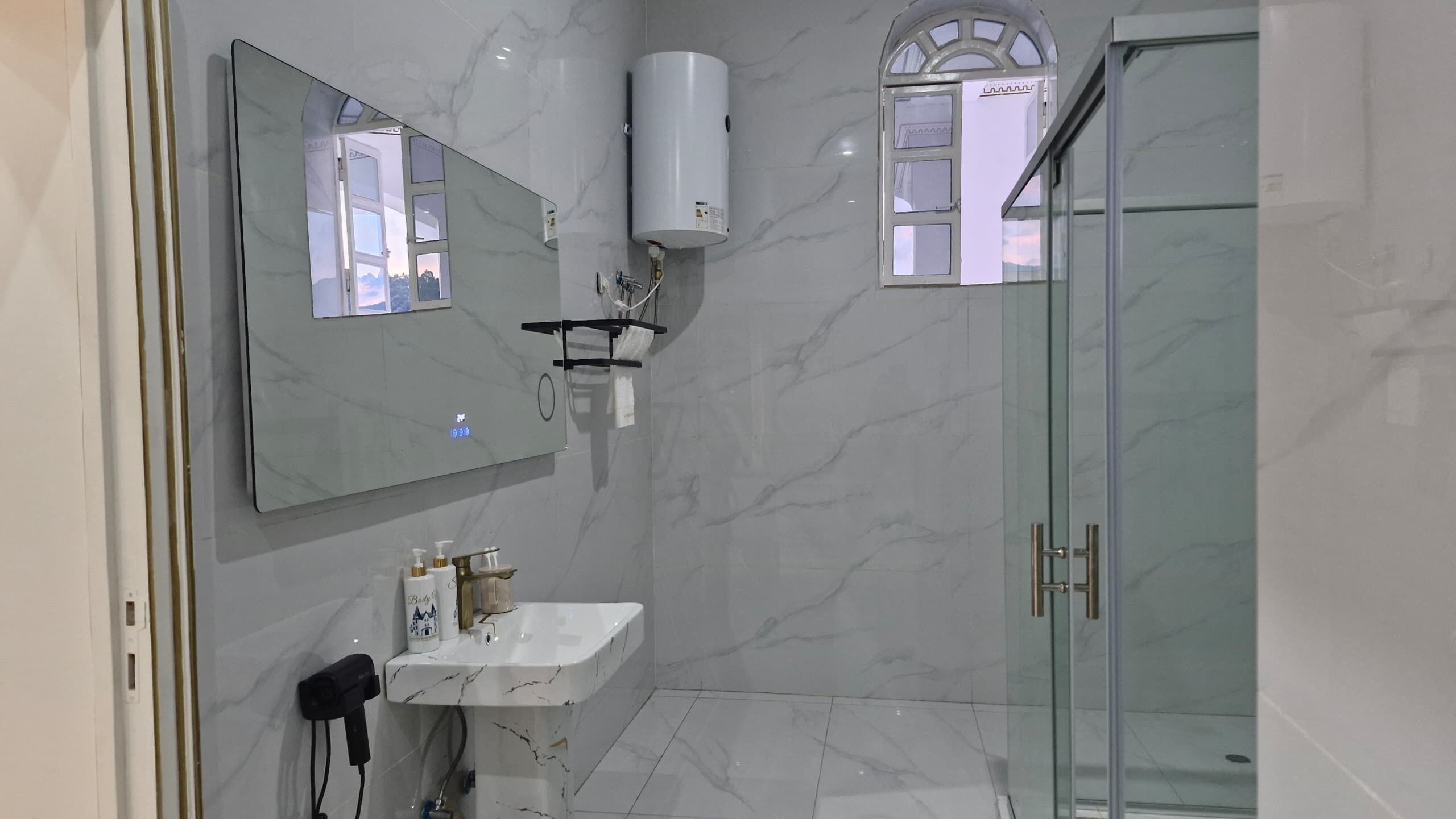 A modern bathroom with a marble floor, a large mirror, a built-in sink, a handheld showerhead, and a glass-enclosed shower.