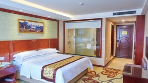 A hotel room with a large bed, a glass-enclosed shower, a wooden headboard, a painting on the wall, and a carpet with a floral pattern. - Vienna hotel shenzhen shajing shangnan  (Shenzhen)