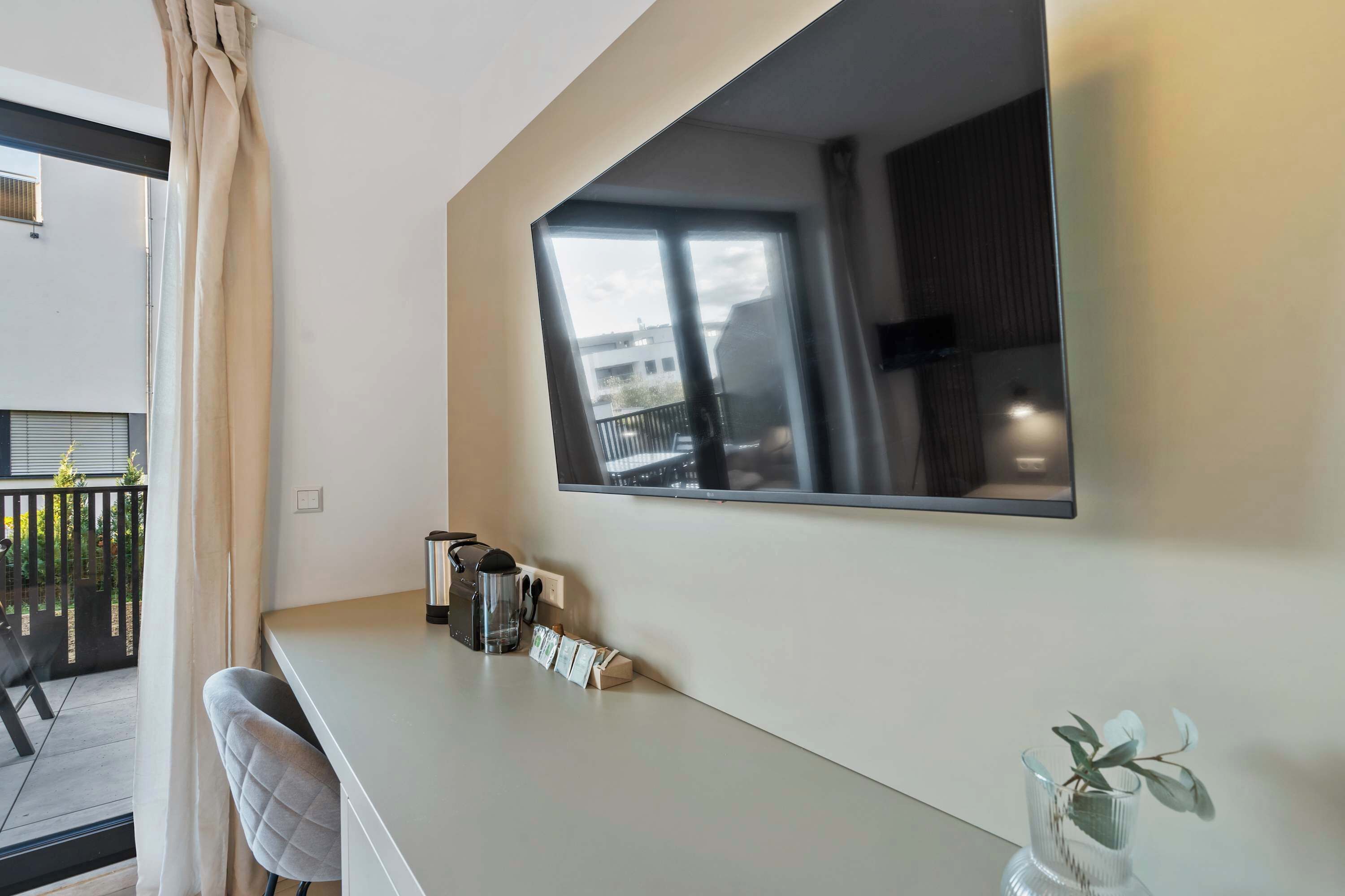 Et moderne hotelværelse med et stort vindue, et fladskærms-tv, et lille bord med elkedel og teposer, en stol og en vase med en plante.